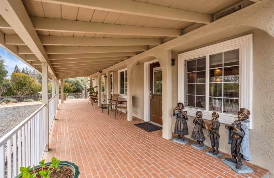 Covered brick front porch with chairs, potted plants, and a row of decorative statues beside a front door and window.