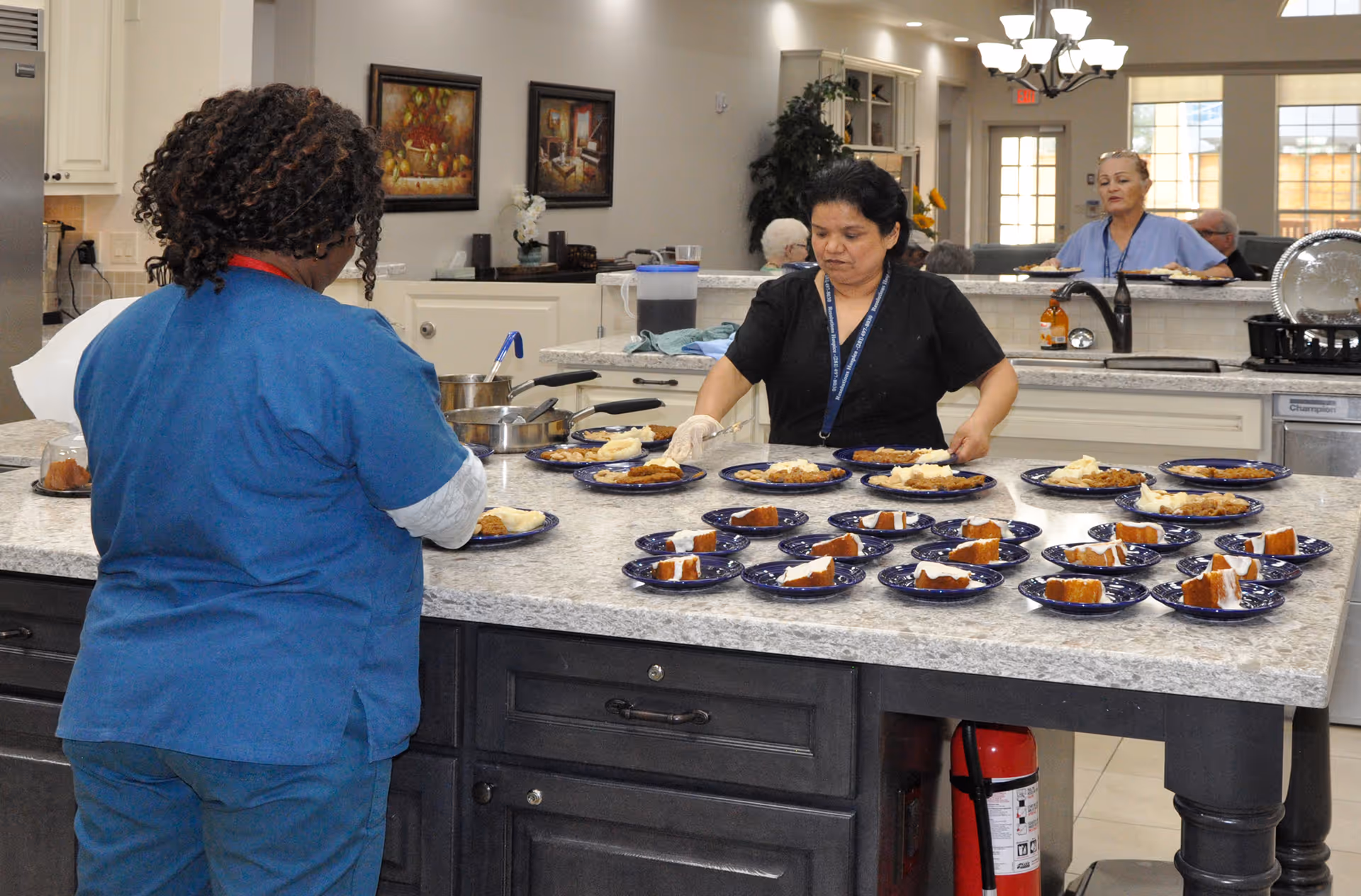 Two staff members in a kitchen area preparing and plating multiple servings of food on a large island countertop. The kitchen has white cabinets, stainless steel appliances, and decorative paintings on the wall. In the background, another staff member is carrying plates, and some elderly residents are seated in a common area.