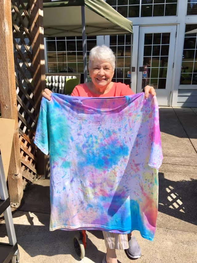 An elderly woman with short white hair is smiling and holding up a large tie-dye t-shirt with vibrant colors including blue, purple, pink, and yellow. She is outdoors on a sunny day, standing on a concrete patio in front of a building with large glass doors and windows. There is a wooden lattice structure to her left and some outdoor furniture visible behind her.