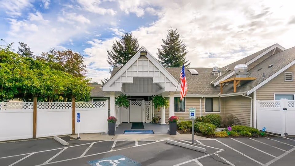 Exterior front entrance of a senior living facility with a covered porch, two large planters with flowers, an American flag, and designated handicapped parking spaces in front. The building has beige siding and white fencing on either side of the entrance.