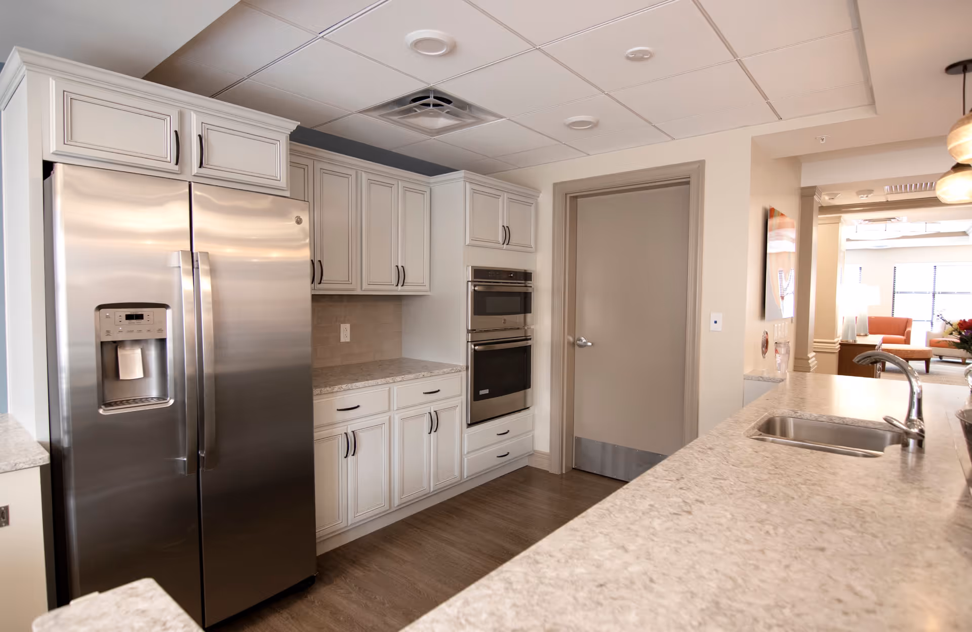 Modern kitchen area with stainless steel refrigerator, built-in double oven, white cabinetry, and a long countertop with a sink. The kitchen opens into a living area with seating and large windows letting in natural light.
