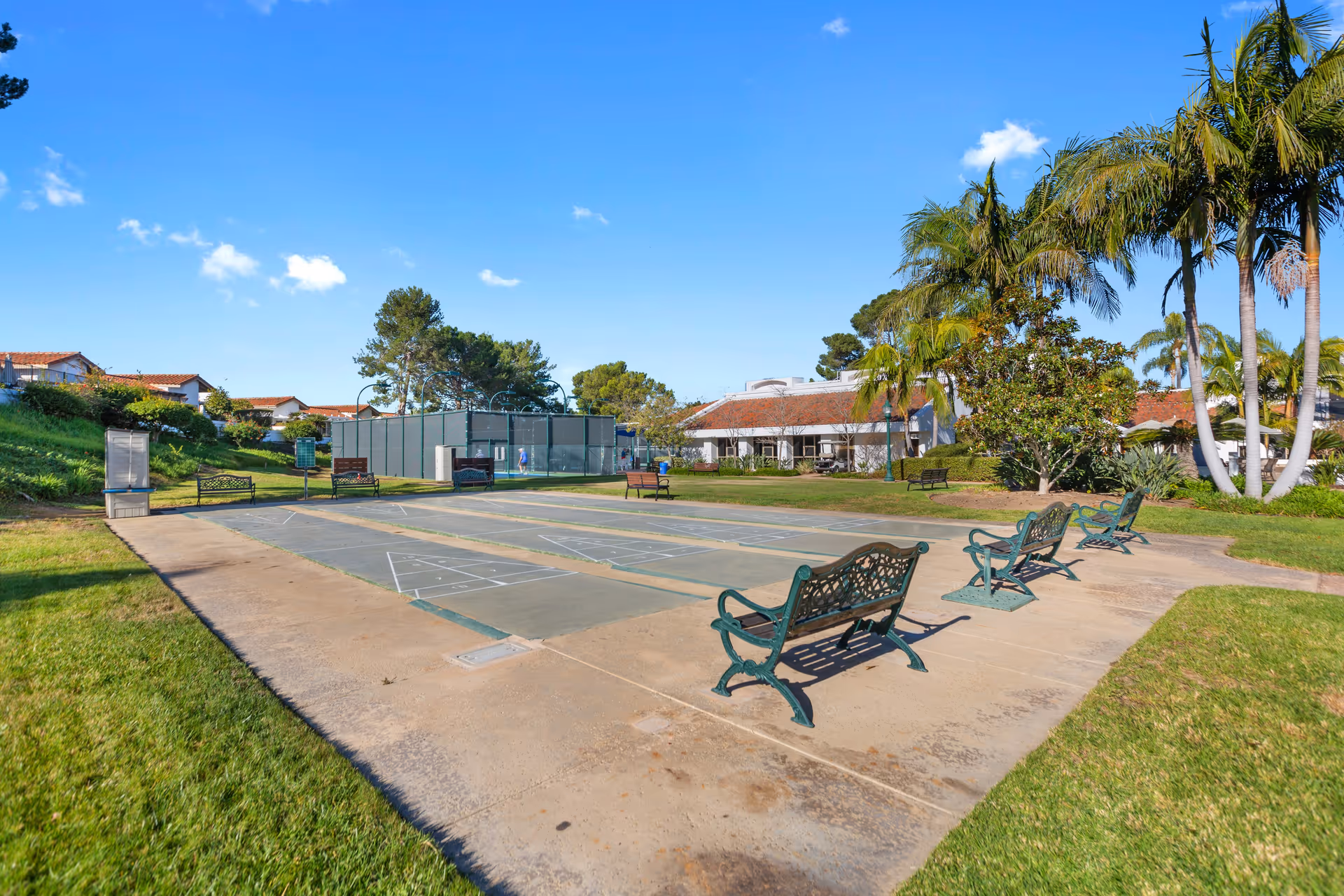 Outdoor shuffleboard courts with green metal benches and palm trees in front of a clubhouse under a blue sky.