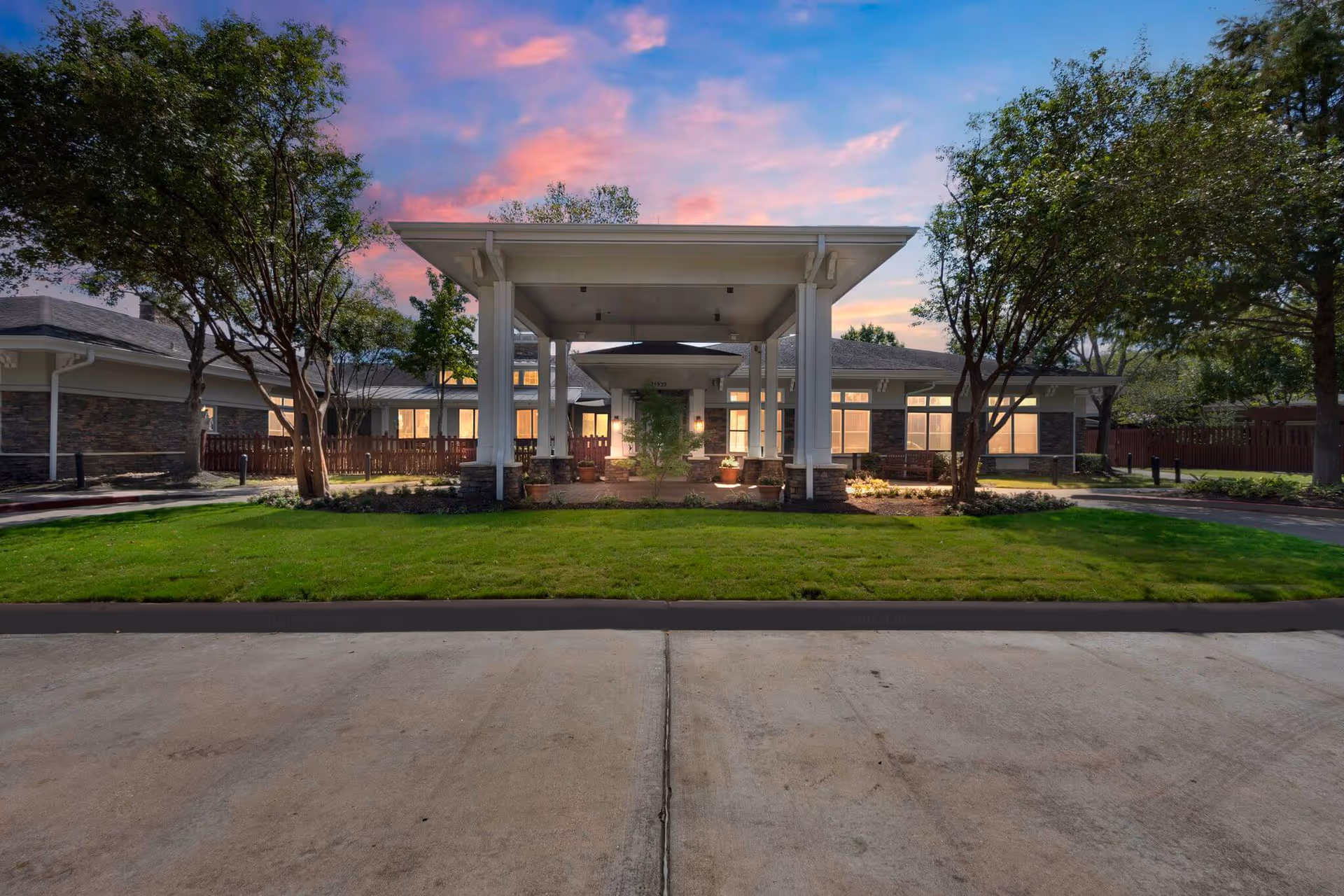 Front entrance of a single-story residential facility with a covered porte-cochère, lit windows, green lawn and trees at dusk.