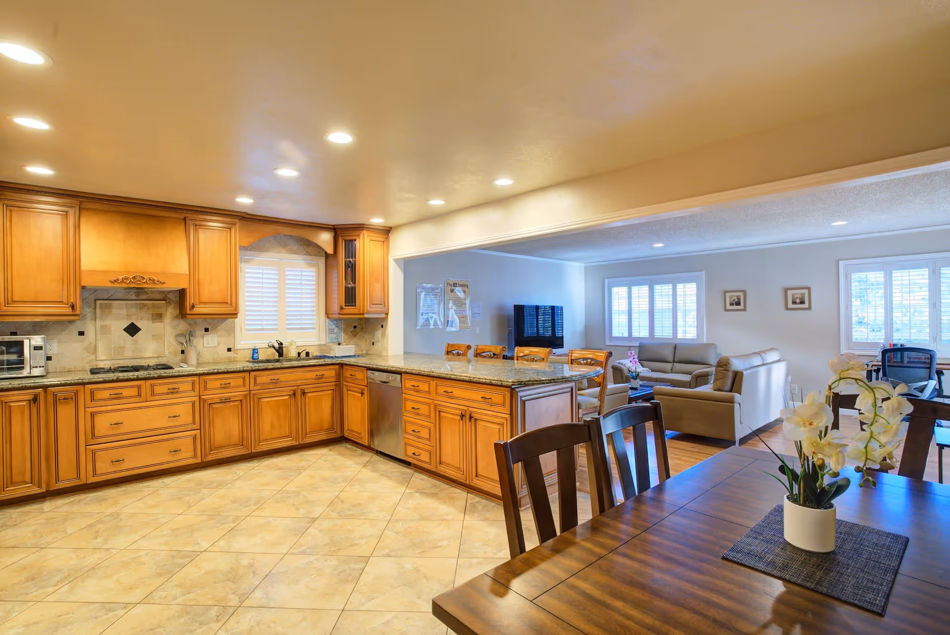 Spacious kitchen with wooden cabinets, granite countertops, and tiled floor. The kitchen opens into a living room area with sofas, a TV, and large windows with white shutters. A wooden dining table with chairs and a white orchid centerpiece is visible in the foreground.