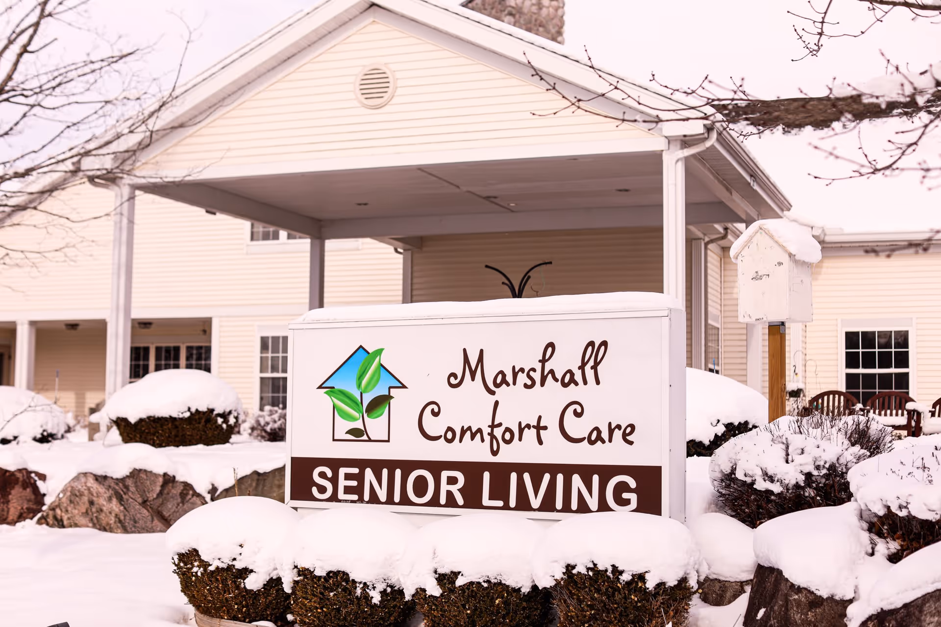 Snow-covered bushes and rocks in front of a sign that reads 'Marshall Comfort Care SENIOR LIVING' with a logo of a house and green leaves, with a beige building and leafless tree branches in the background.