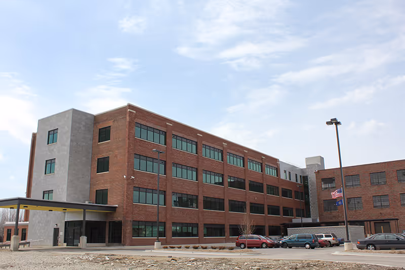 Exterior view of a large four-story brick building with many windows, a covered entrance, several parked cars, and two flagpoles with flags flying, under a partly cloudy sky.