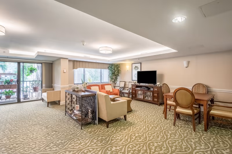 Spacious senior living common room with comfortable seating, a TV console, dining table, and sliding glass doors to a balcony.