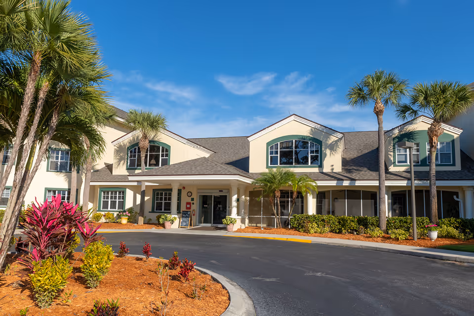 Exterior view of a senior living facility building with a curved driveway, palm trees, and landscaped garden beds under a clear blue sky.