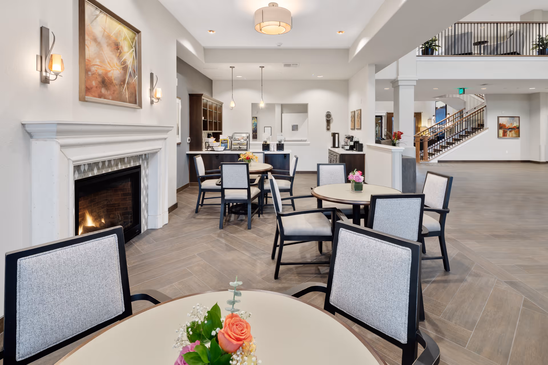 A spacious and well-lit common area in a senior living facility featuring round tables with cushioned chairs, small floral centerpieces on each table, a lit fireplace with a painting above it, and a counter area with coffee machines and pendant lights. In the background, there is a staircase leading to an upper floor and some framed artwork on the walls.