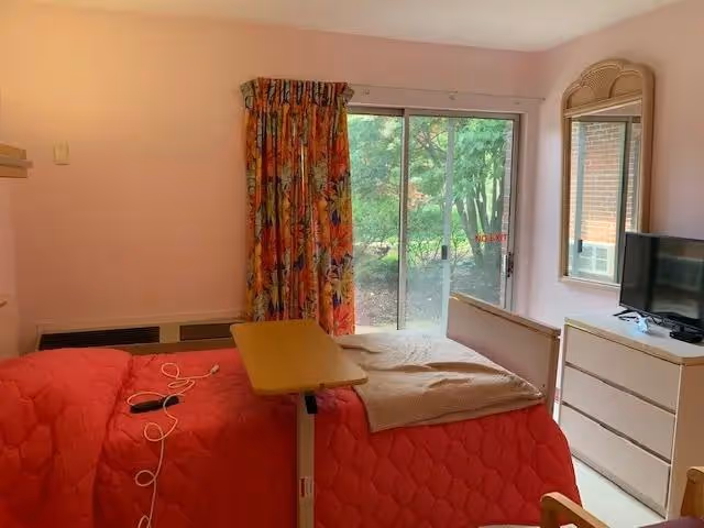A small bedroom with a bed covered in a red quilt and a beige blanket. There is a wooden overbed table on the bed, a dresser with a flat-screen TV on top, and a wall-mounted mirror above the dresser. A sliding glass door with floral curtains leads to an outdoor area with greenery visible outside.