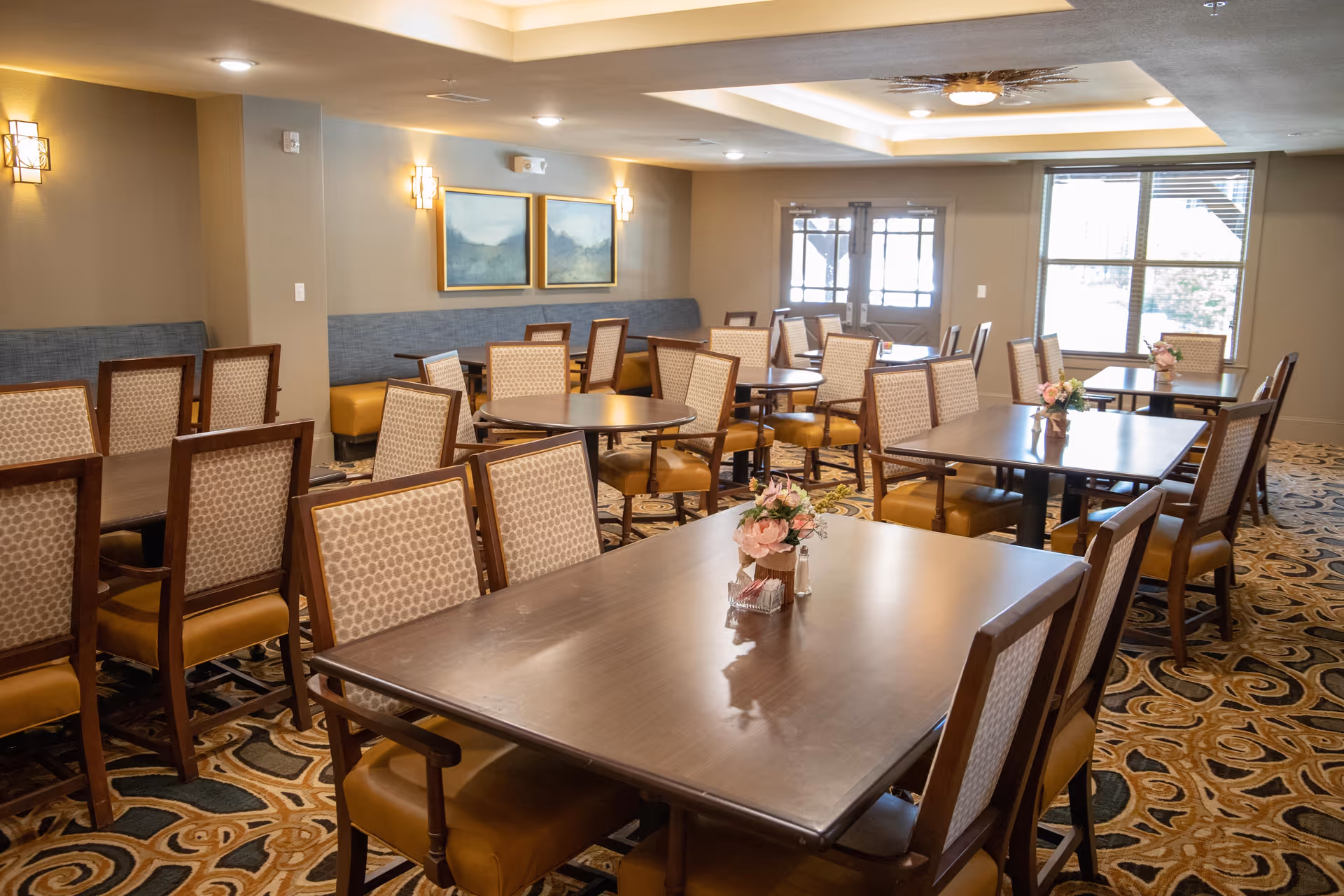 A dining room with multiple wooden tables and cushioned chairs arranged neatly. Each table has a small floral centerpiece. The room has patterned carpet flooring, beige walls with framed artwork, and wall-mounted lights. Large windows and glass doors allow natural light to enter the space.