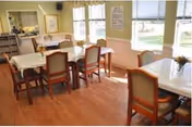 A bright dining room with several tables covered with white tablecloths and surrounded by wooden chairs with cushioned seats. Large windows allow natural light to fill the room, and there are framed pictures and a mirror on the walls. The floor is wooden, and the overall atmosphere is clean and inviting.