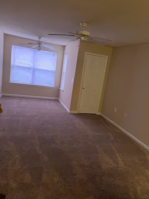 Empty carpeted living area with two ceiling fans, a large window with blinds, and a closed door.