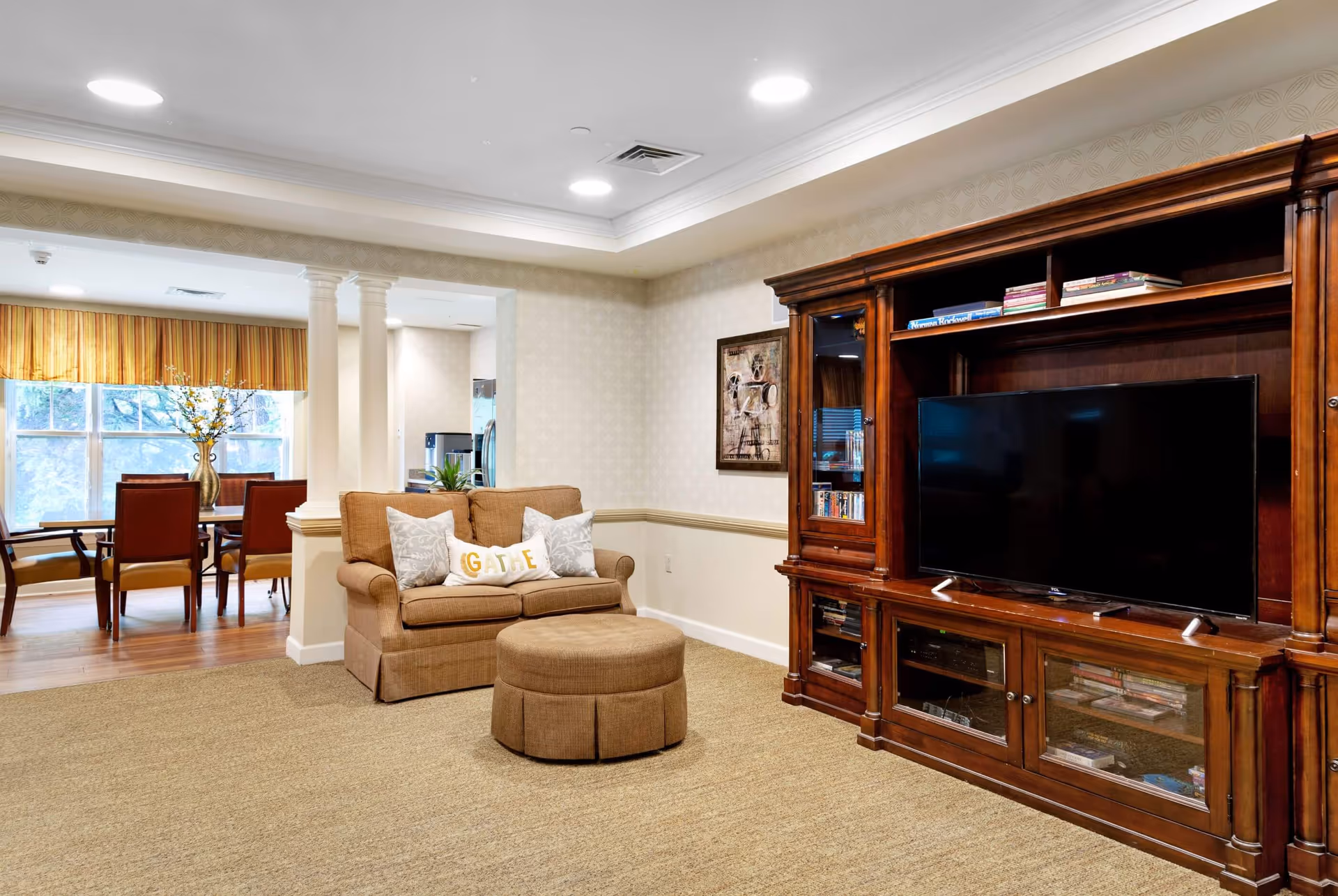 A cozy living room area with a brown loveseat and matching ottoman, a large wooden entertainment center with a flat-screen TV, and a dining area with a table and chairs visible in the background near large windows with striped valances.