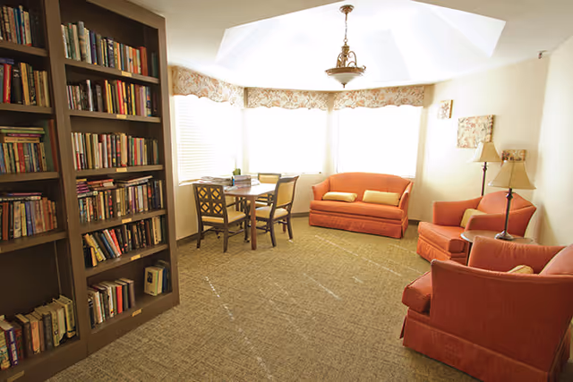 A cozy living room area with a large bookshelf filled with books on the left, a small wooden table with four chairs near the windows, and three orange upholstered seating pieces including a loveseat and two armchairs. The room is well-lit with natural light coming through the windows, and there are two floor lamps and a ceiling light fixture.