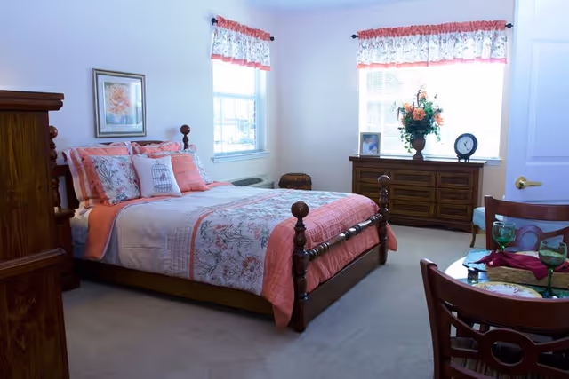 A bedroom with a wooden bed frame featuring floral and coral bedding, two windows with floral valances, a wooden dresser with a vase of flowers, a clock, and a framed photo, and a small dining table with chairs and place settings.