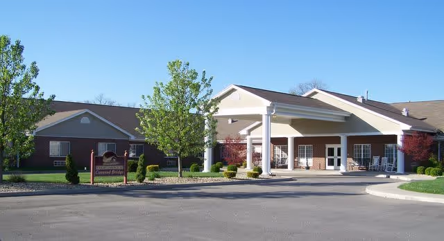 Exterior view of Covered Bridge Health Campus showing a single-story brick building with a covered entrance supported by white columns. There are small trees and shrubs in landscaped areas near the entrance and a paved driveway in front.