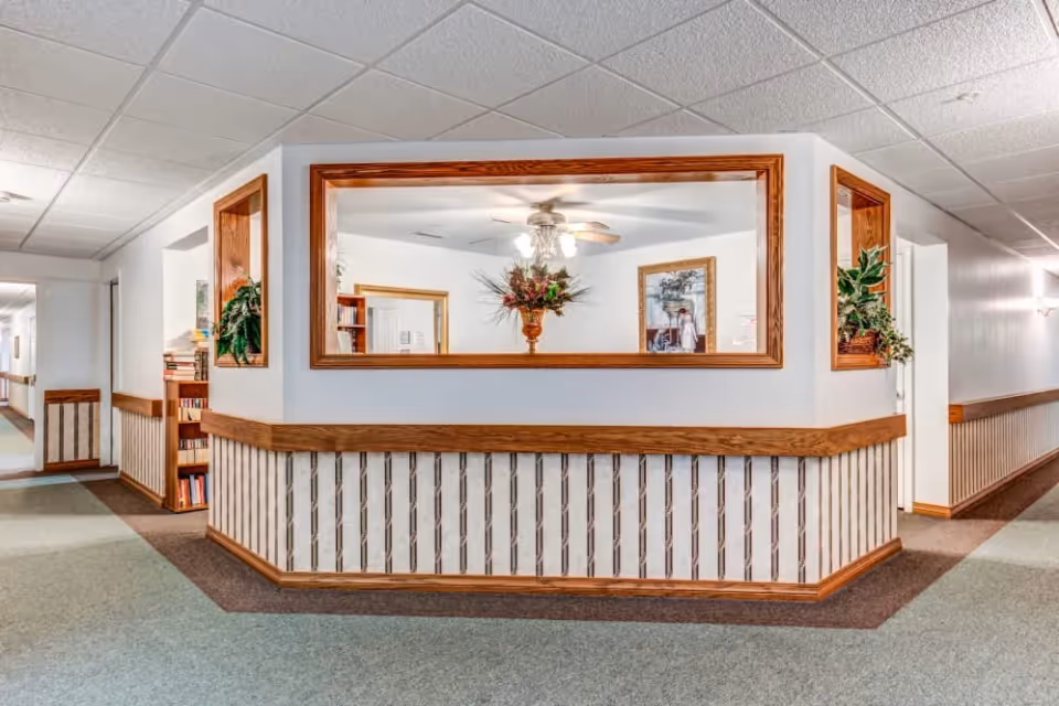 Interior hallway reception desk with wood-trimmed pass-through window, plants, and framed artwork.