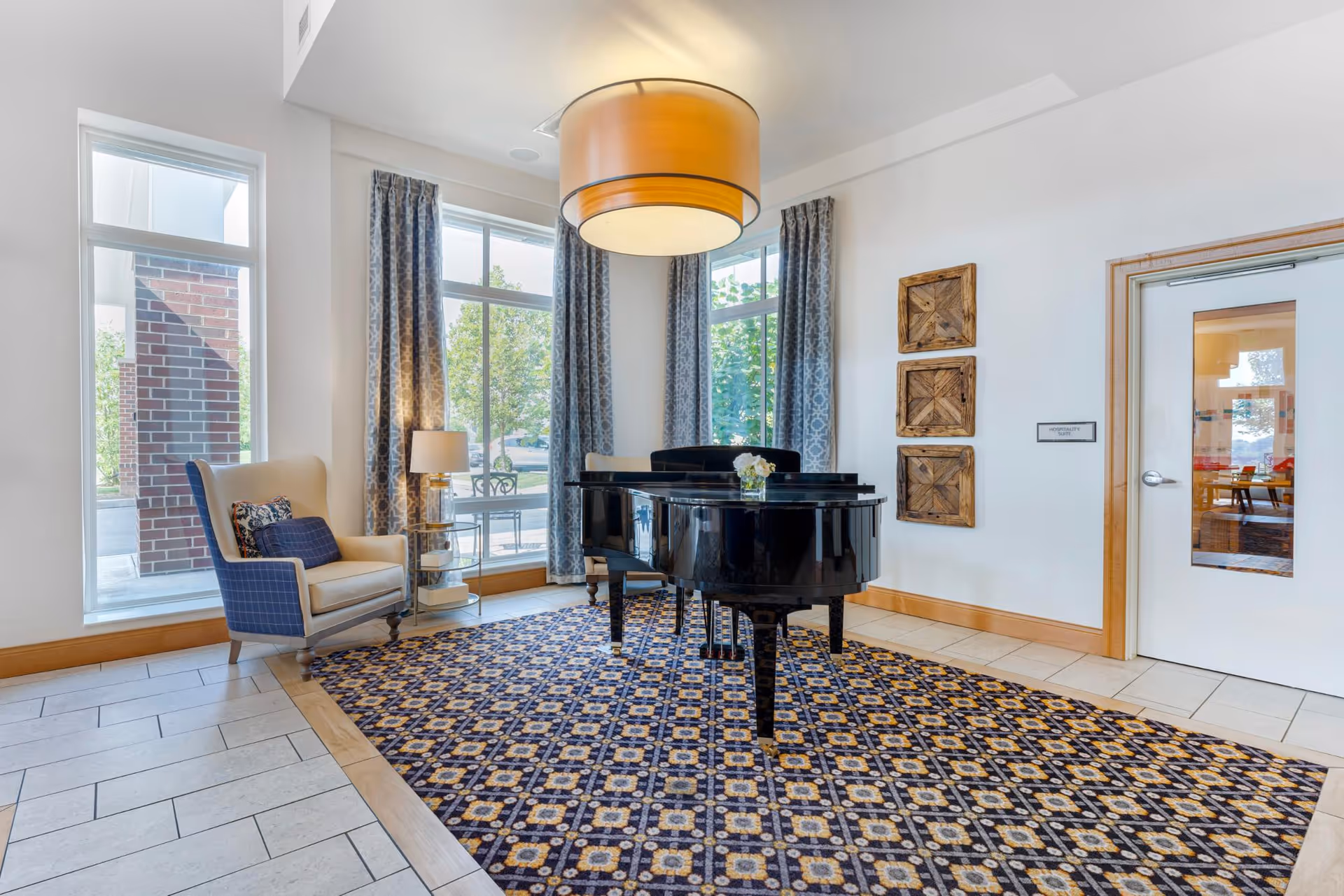 A bright room with large windows and patterned curtains featuring a black grand piano on a decorative carpet. To the left, there is a beige armchair with a blue cushion and a small side table with a lamp. The walls are white with three wooden square wall decorations and a door with a window on the right side.