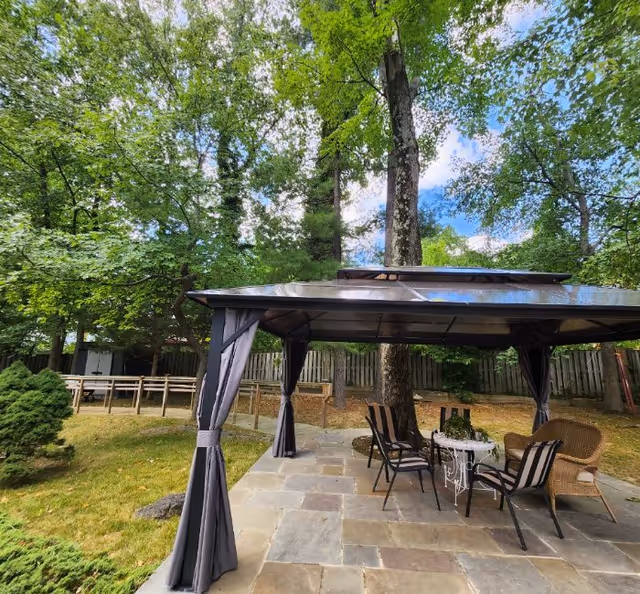 Stone patio with a covered gazebo and outdoor seating set among trees and a wooden fence.