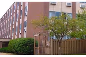 Exterior view of a multi-story brick building with several windows, surrounded by green bushes and a tree in front of a wooden fence.