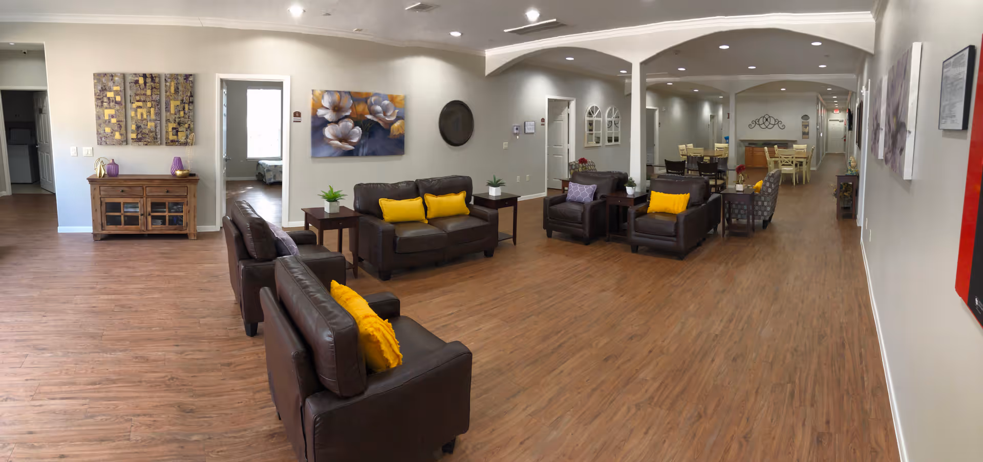 A spacious and well-lit common area in an assisted living facility featuring brown leather sofas and armchairs with yellow and purple cushions, wooden side tables with small plants, and wall art including floral paintings and abstract pieces. The room has wood flooring and leads to a dining area with tables and chairs in the background.