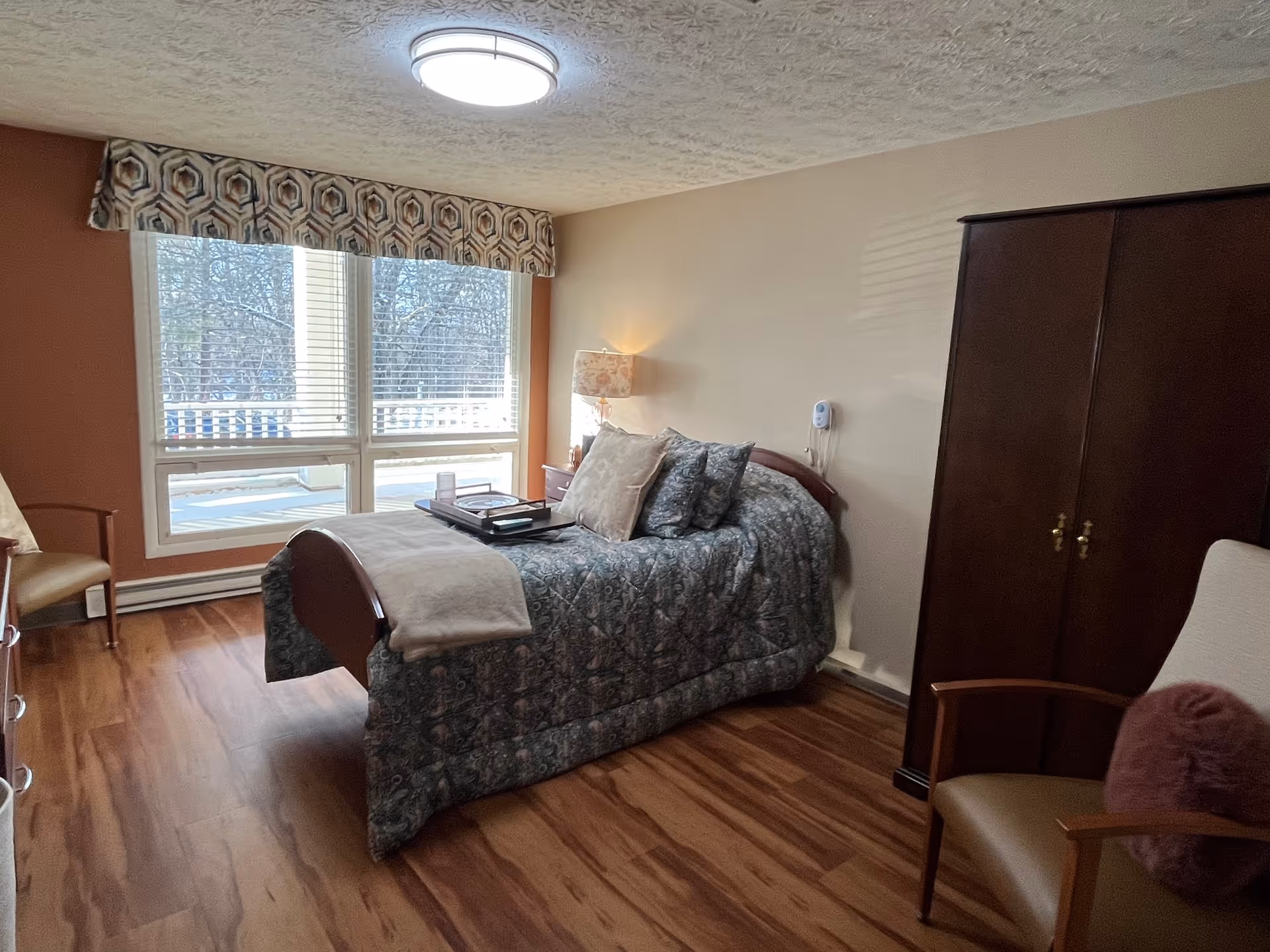 Sunlit furnished bedroom with a made bed, bedside lamp, large window with valance, wooden wardrobe and seating.