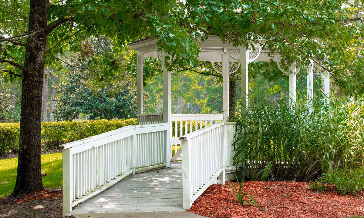 A white wooden gazebo with a ramp surrounded by green trees, bushes, and landscaped garden beds with red mulch at The Gables of Tallahassee.