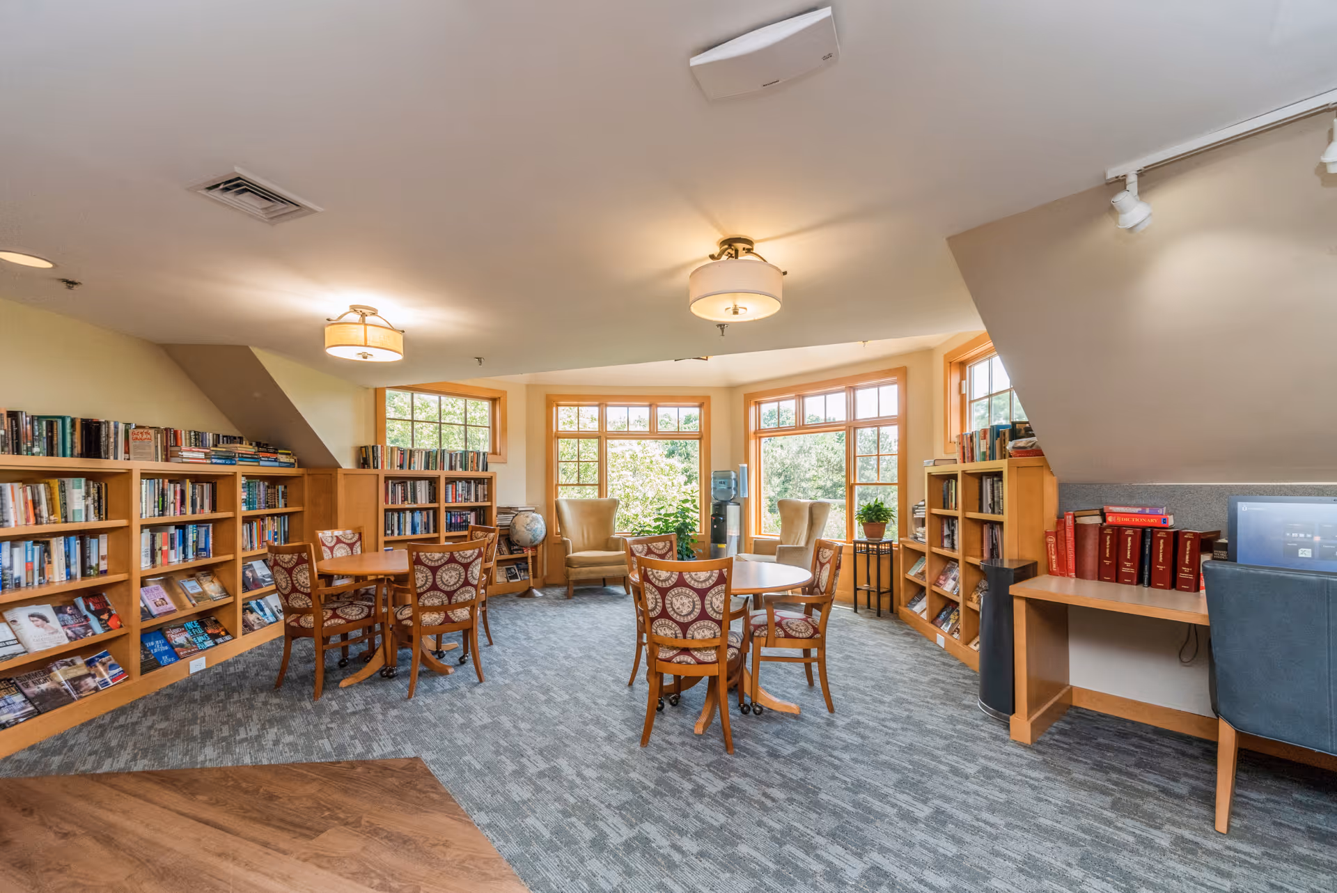 A bright and cozy library or reading room with large windows letting in natural light. The room features wooden bookshelves filled with books, round tables with patterned chairs, two beige armchairs near the windows, a globe, a water cooler, and a desk with a computer and blue chair. The carpet is gray with a subtle pattern, and the ceiling has modern light fixtures.