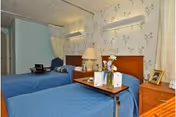 A nursing facility patient room with two twin beds in blue bedspreads, a shared nightstand with a lamp and flowers, and floral wallpaper.
