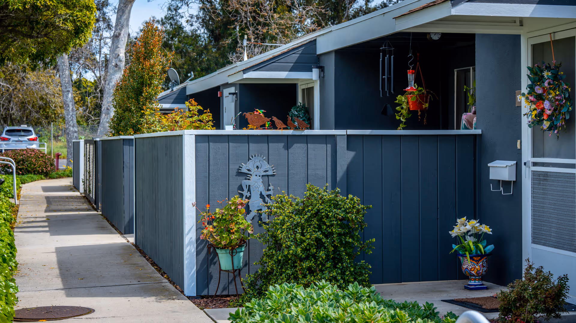Exterior view of a residential unit at Ventura Townehouse featuring a gray wooden fence with decorative metal art and potted plants. A concrete walkway runs alongside the fence, leading to the entrance door adorned with a colorful floral wreath. Trees and greenery surround the area, and a parked car is visible in the background.