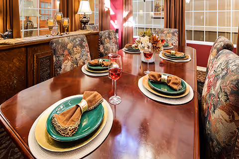 Polished dining table in a formal dining room set with green plates, brown napkins, wine glasses, and floral upholstered chairs.