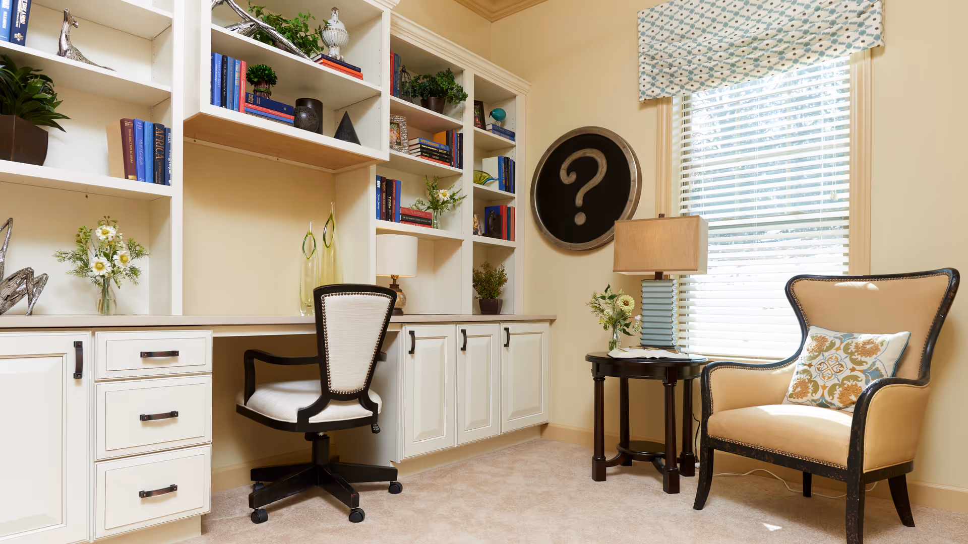 Bright home office nook with built-in white shelving and desk, a swivel desk chair, a beige armchair by a window, and a side table with a lamp.