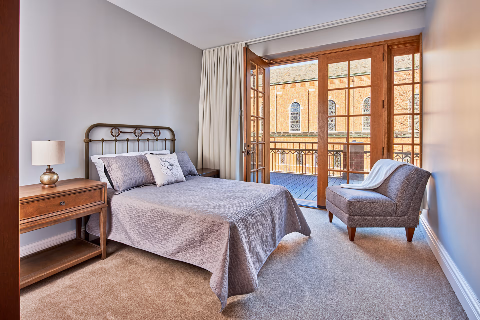 A bedroom with a metal framed bed covered with a gray quilt and pillows, a wooden nightstand with a lamp, a gray upholstered chair with a white throw blanket, and large wooden-framed glass doors leading to a balcony with a view of a brick building with arched windows.