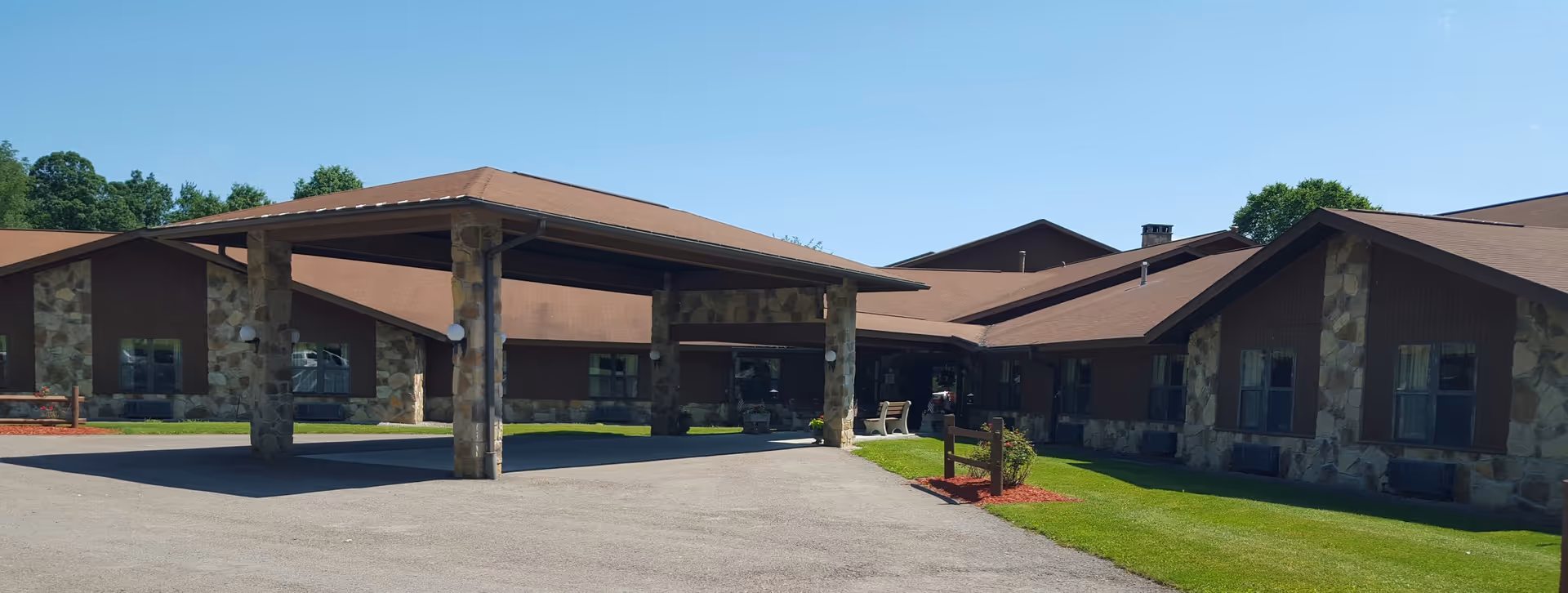 Exterior view of Siemon's Heritage Personal Care Center building with stone pillars and brown roof under a clear blue sky. The entrance has a covered driveway with stone columns and there is green grass and some landscaping around the building.
