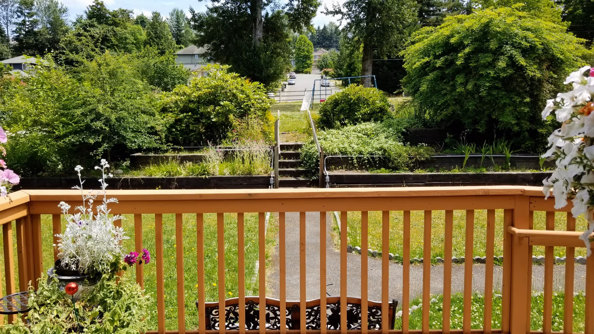 View from a wooden deck railing overlooking a grassy yard with potted flowers, a walkway and steps leading up through terraced landscaping to a street.