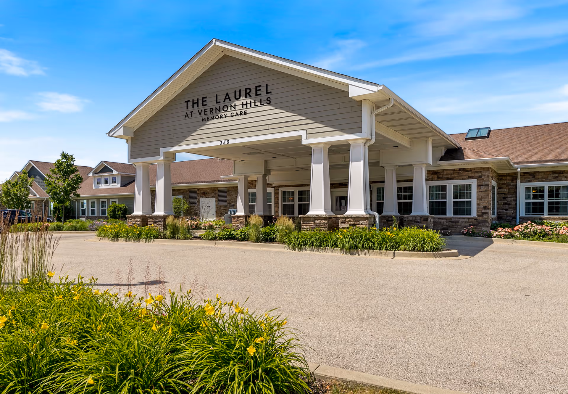 Exterior front view of The Laurel at Vernon Hills Memory Care facility with a covered entrance supported by white columns, surrounded by landscaping with green plants and yellow flowers under a blue sky.