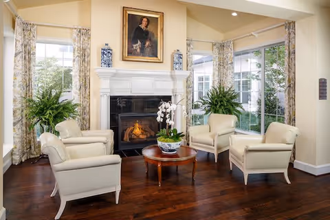 Bright sitting room with four cream armchairs arranged around a round wooden coffee table facing a lit fireplace, flanked by large windows and potted plants.