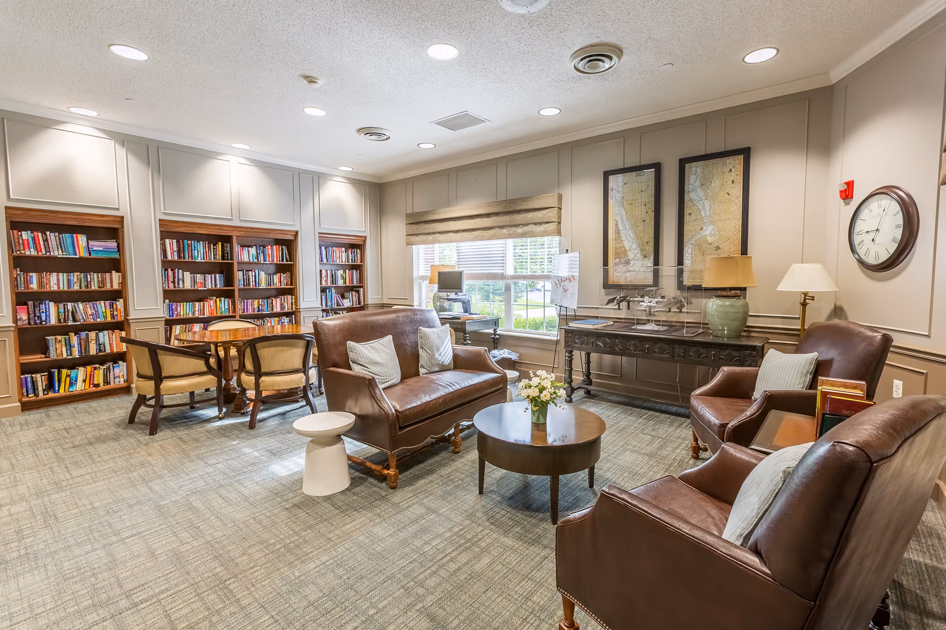 A cozy living room area in a senior living facility featuring leather armchairs and a loveseat with cushions, a round wooden coffee table with a small flower arrangement, a side table with books, and a wooden table with four chairs near built-in bookshelves filled with books. The room has beige paneled walls, two framed maps, a large window with a beige roman shade, and two table lamps. A wall clock is mounted on the right wall.