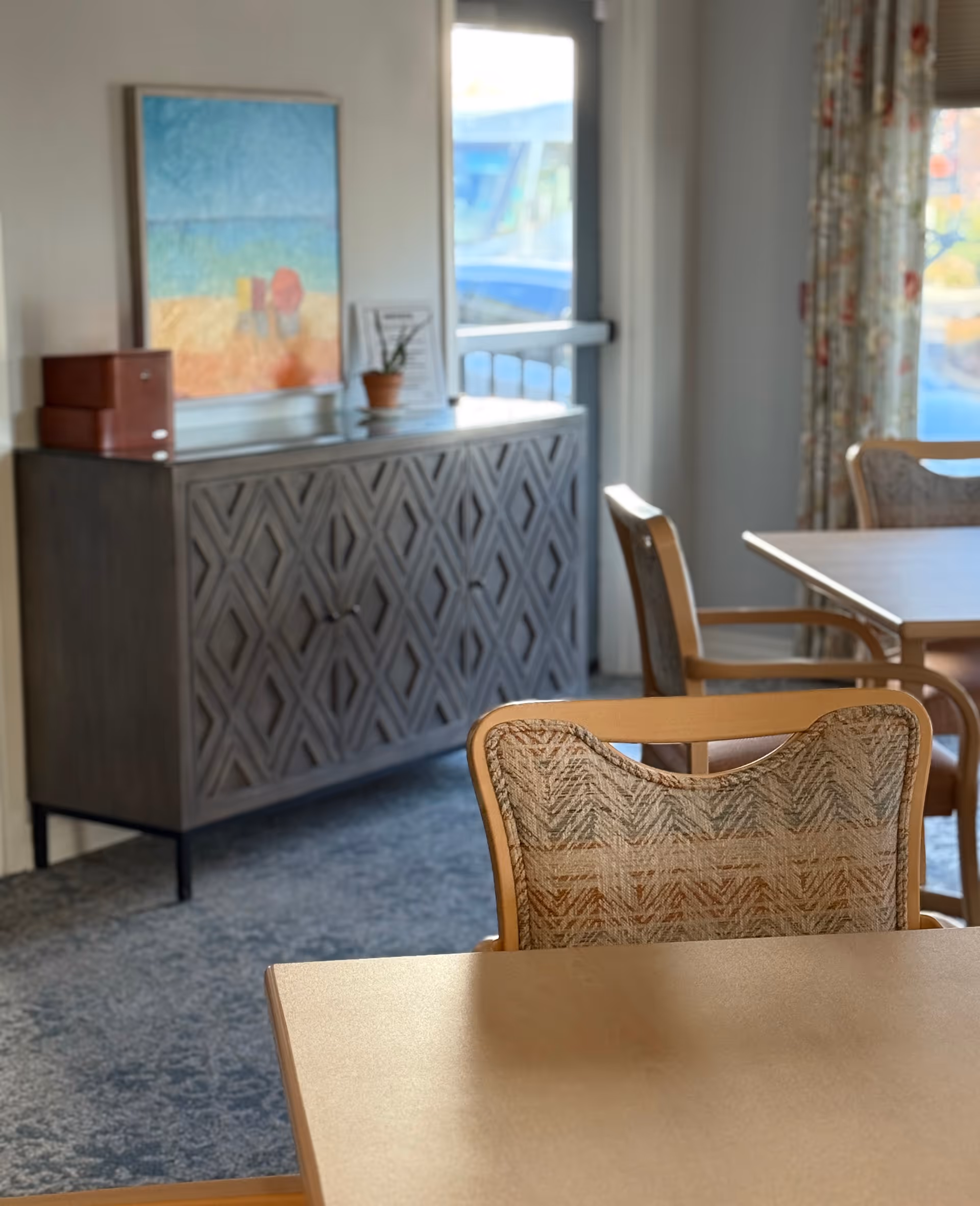 A bright dining area with wooden tables and cushioned chairs featuring patterned upholstery. In the background, there is a decorative cabinet with a geometric design, a small potted plant, a framed picture, and a colorful abstract painting on the wall. Sunlight streams in through a window with floral curtains.