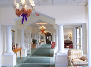 A bright and spacious hallway in a senior living facility with white columns and archways, green carpet with patterned borders, chandeliers decorated with autumn-themed wreaths, and seating areas with upholstered chairs. The hallway leads to other rooms with large windows allowing natural light.