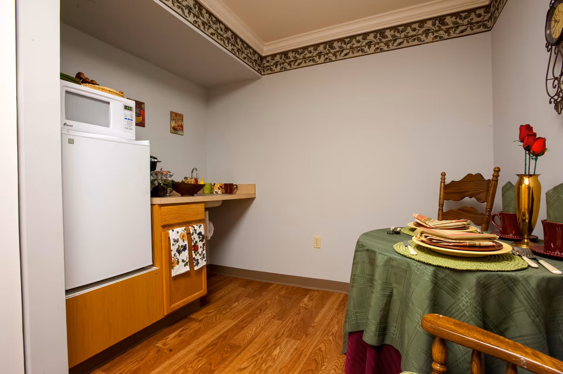 A small dining area with a round table covered with a green tablecloth, set with plates, napkins, and utensils. There are two wooden chairs around the table. To the left, there is a compact kitchen area with a white microwave on top of a white refrigerator, wooden cabinets below, and a countertop with various items. The room has hardwood flooring and a decorative border near the ceiling.