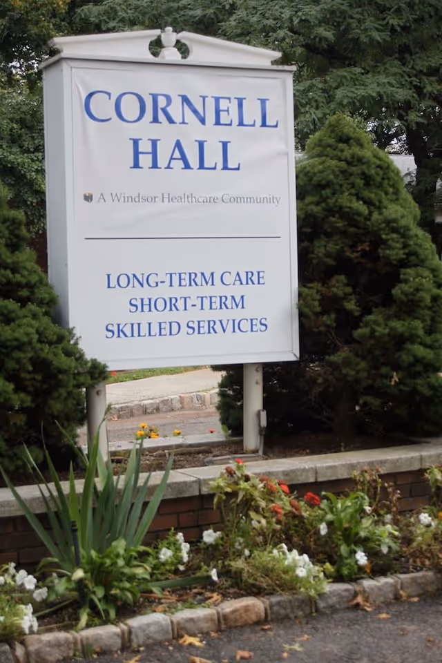 Outdoor sign for Cornell Hall, a Windsor Healthcare Community, offering long-term care, short-term, and skilled services, surrounded by greenery and flowers.