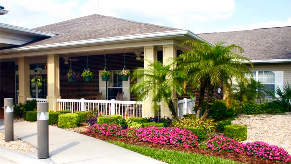 Front entrance of a senior living facility with a covered porch, hanging flower baskets, palm trees, and colorful landscaped flower beds.