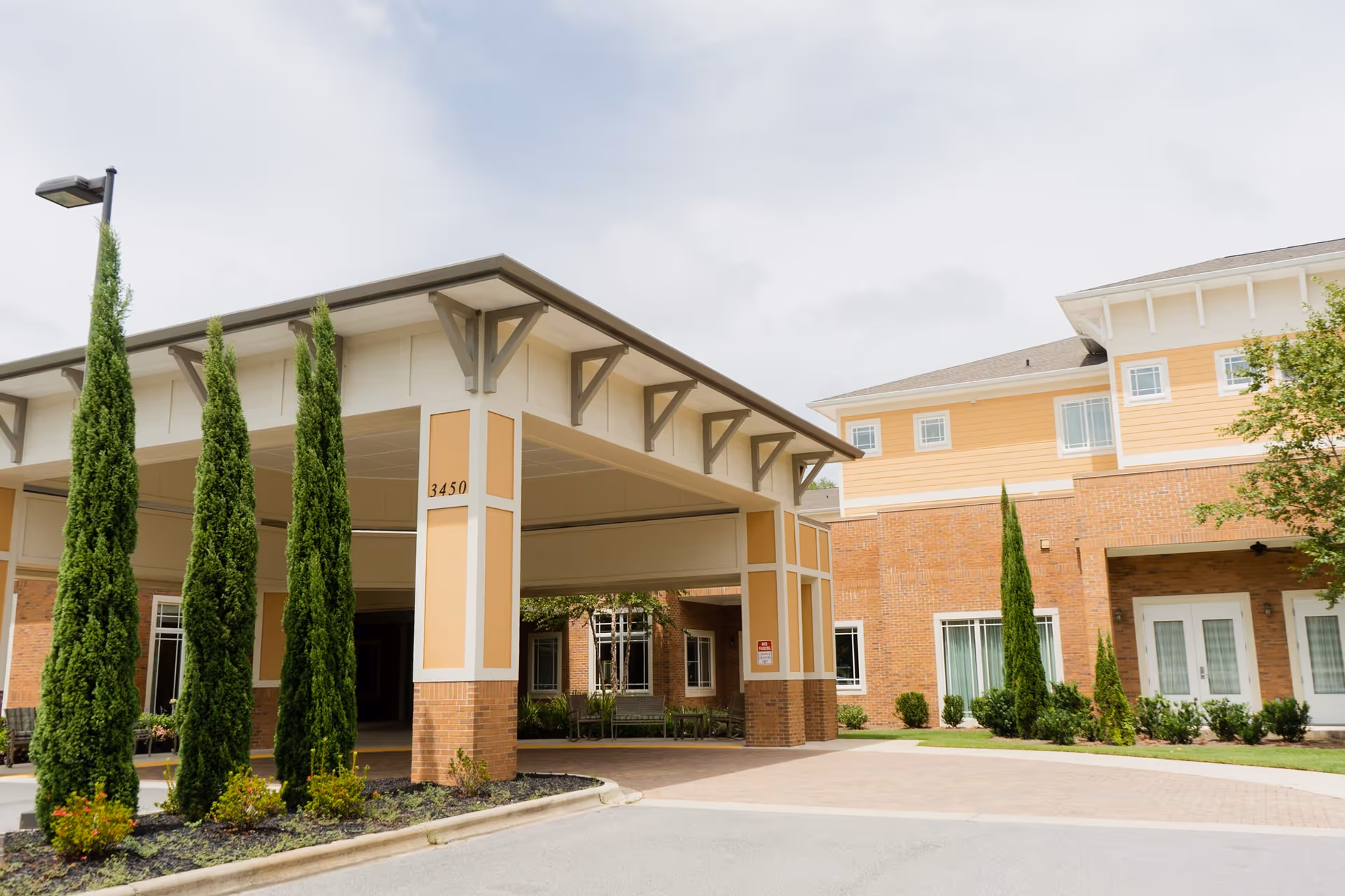 Exterior view of Summer Vista Assisted Living facility showing the entrance with a covered driveway supported by columns, surrounded by neatly trimmed tall green shrubs and a brick building with multiple windows.
