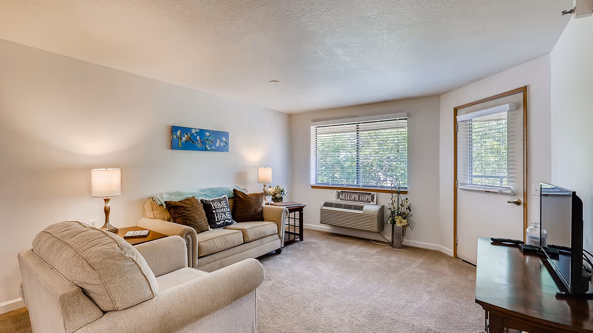 Bright living room with beige sofas and armchair, lamps, a TV on a stand, a large window with blinds and a door to the outside.
