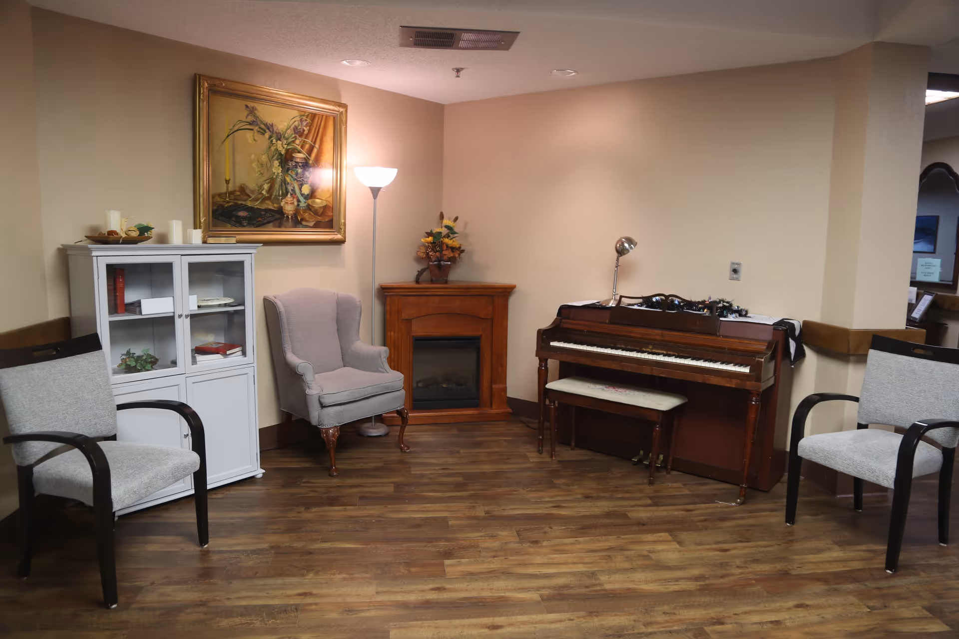 A cozy corner of a room featuring a wooden upright piano with a cushioned bench, a gray upholstered armchair, two gray chairs with black armrests, a white cabinet with glass doors containing books and decorative items, a wooden electric fireplace, a floor lamp, and a framed painting of flowers on the wall.