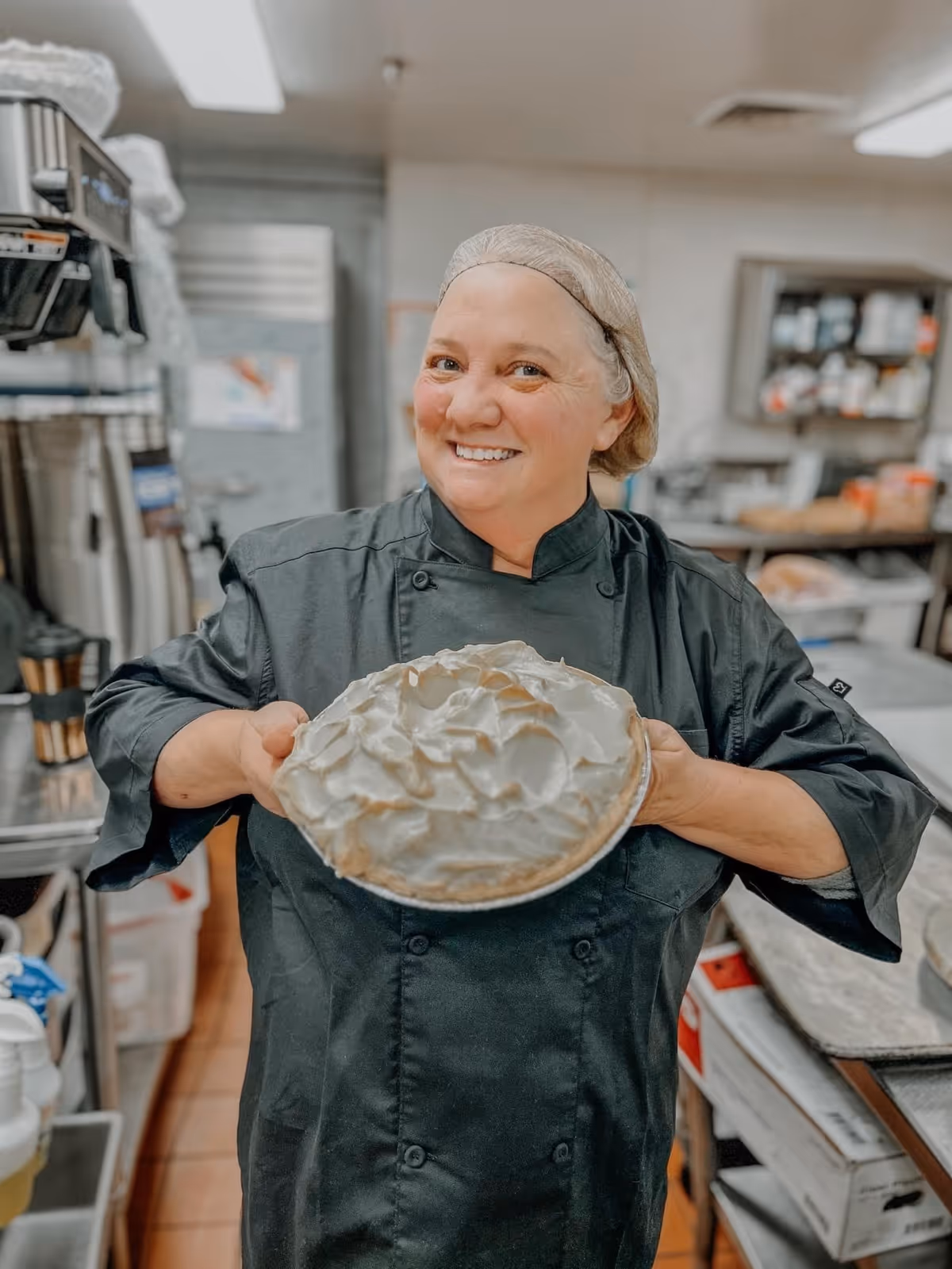 A smiling chef wearing a black uniform and hairnet holds a freshly baked pie with a golden meringue topping in a commercial kitchen.