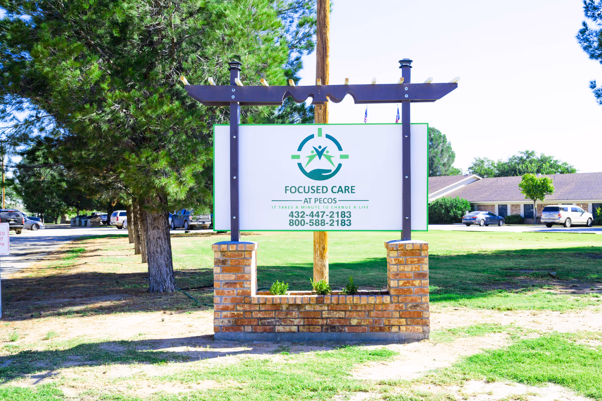 A large outdoor sign for Focused Care at Pecos, mounted on a brick and wood frame, with trees, grass, parked cars, and a building in the background under a clear sky.
