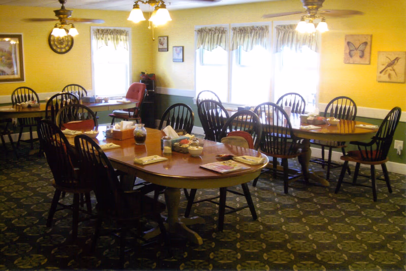 A dining room with several wooden tables and chairs arranged neatly. The tables have napkins, condiments, and small bowls placed on them. The room has yellow walls with green wainscoting, patterned carpet, and windows with valance curtains letting in natural light. Ceiling fans with lights hang from the ceiling, and framed pictures decorate the walls.